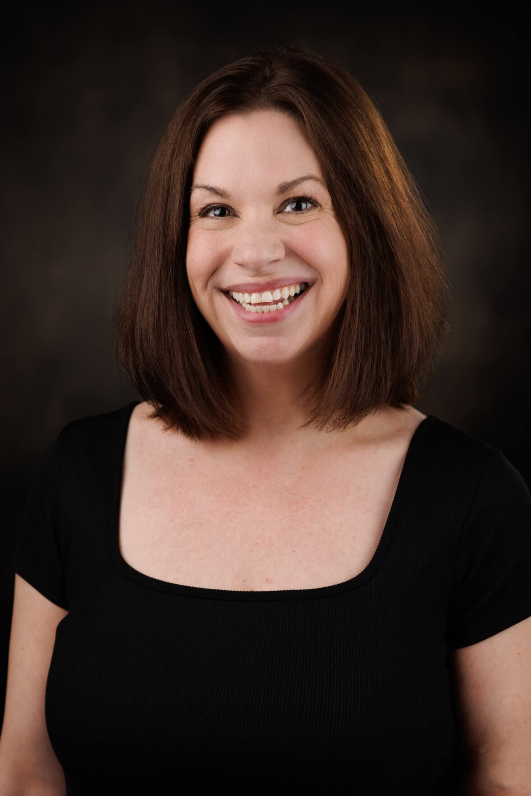 white woman with brown hair wearing a black top, smiling, in front of a brown background