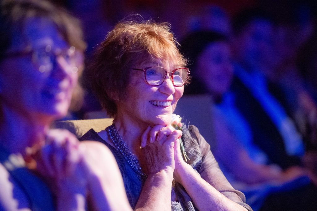 photo of a woman with red hair clasping her hands under her chin in a darm theater. She is smiling.