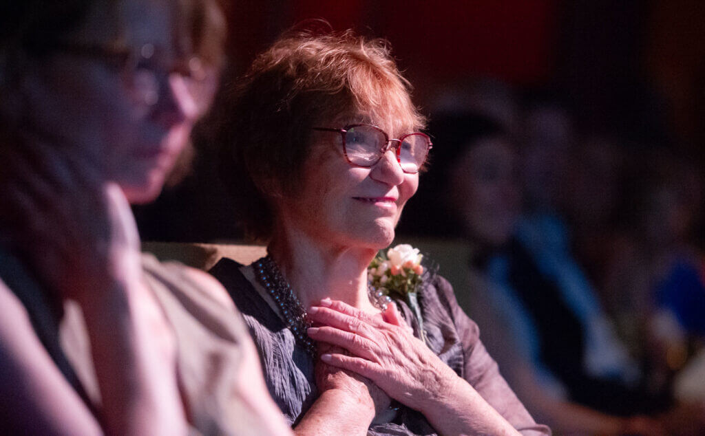 a woman with short red hair and glasses holds her hands to her heart and smiles. She is seated in a theater.