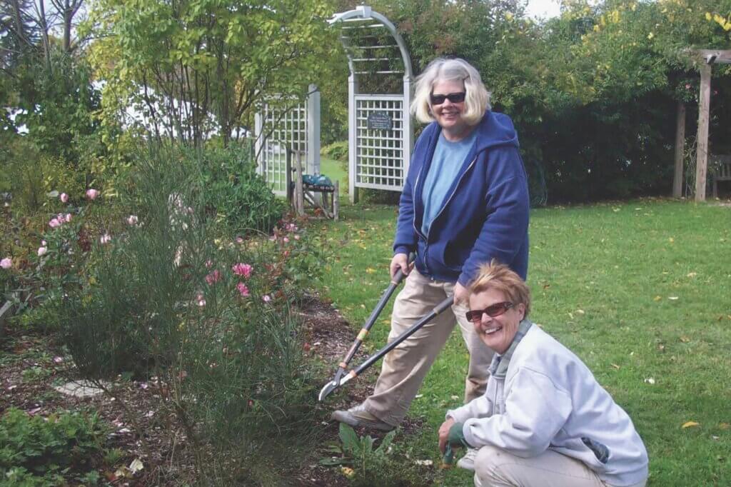 two smiling women gardening. One is standing and the other is kneeling near some shrubbery.