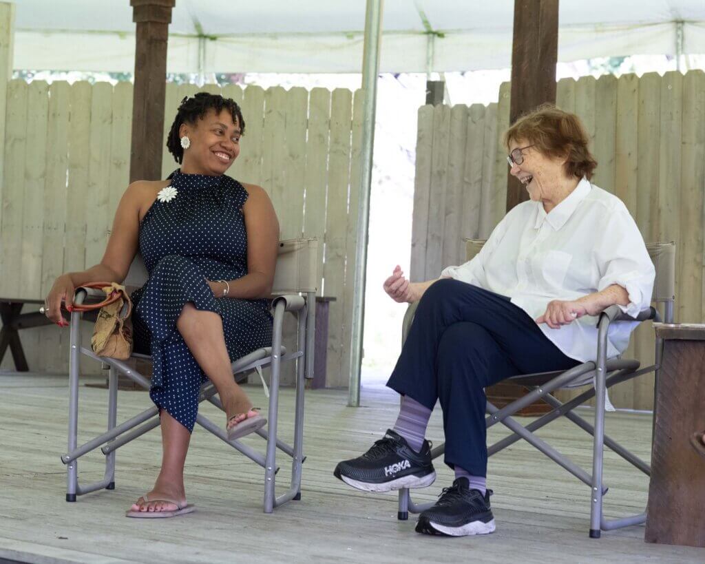 two women sit on an outdoor stage in folding chairs. one is black and one is white. They are looking at each other smiling