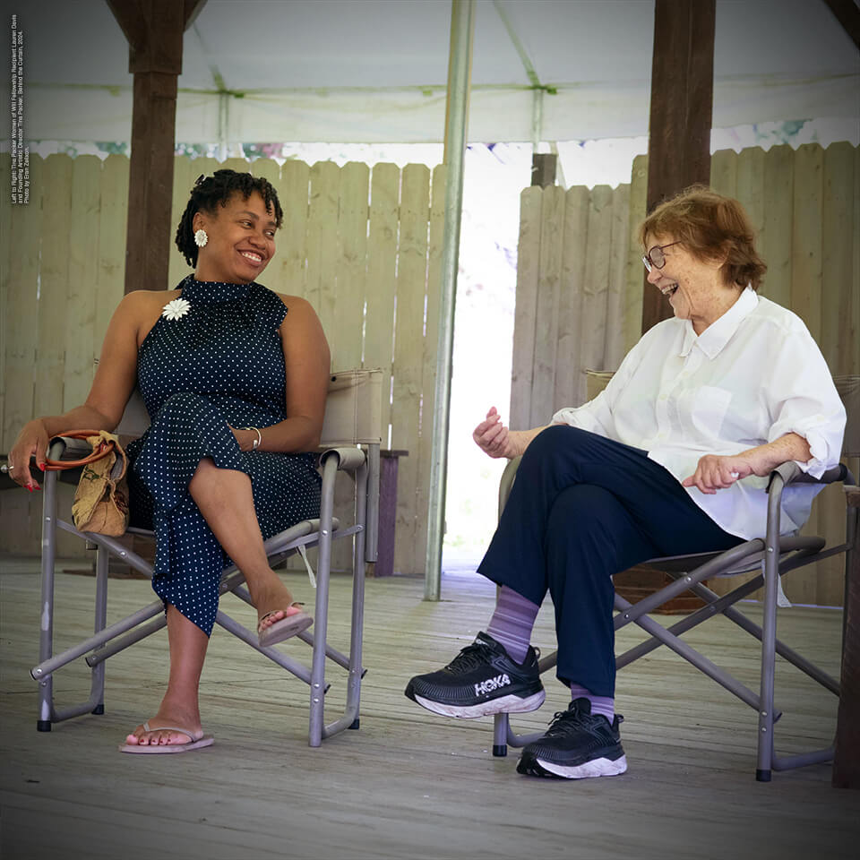 two women sitting on a stage looking at each other. They are both smiling