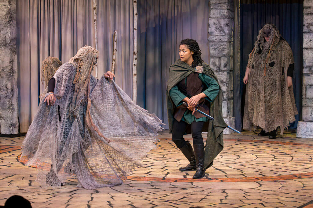 A woman at left dressed in gray gauze faces a woman in a green cape. Another woman stands at right in the same gray gauzy costume. They are performing in a staged production of Macbeth