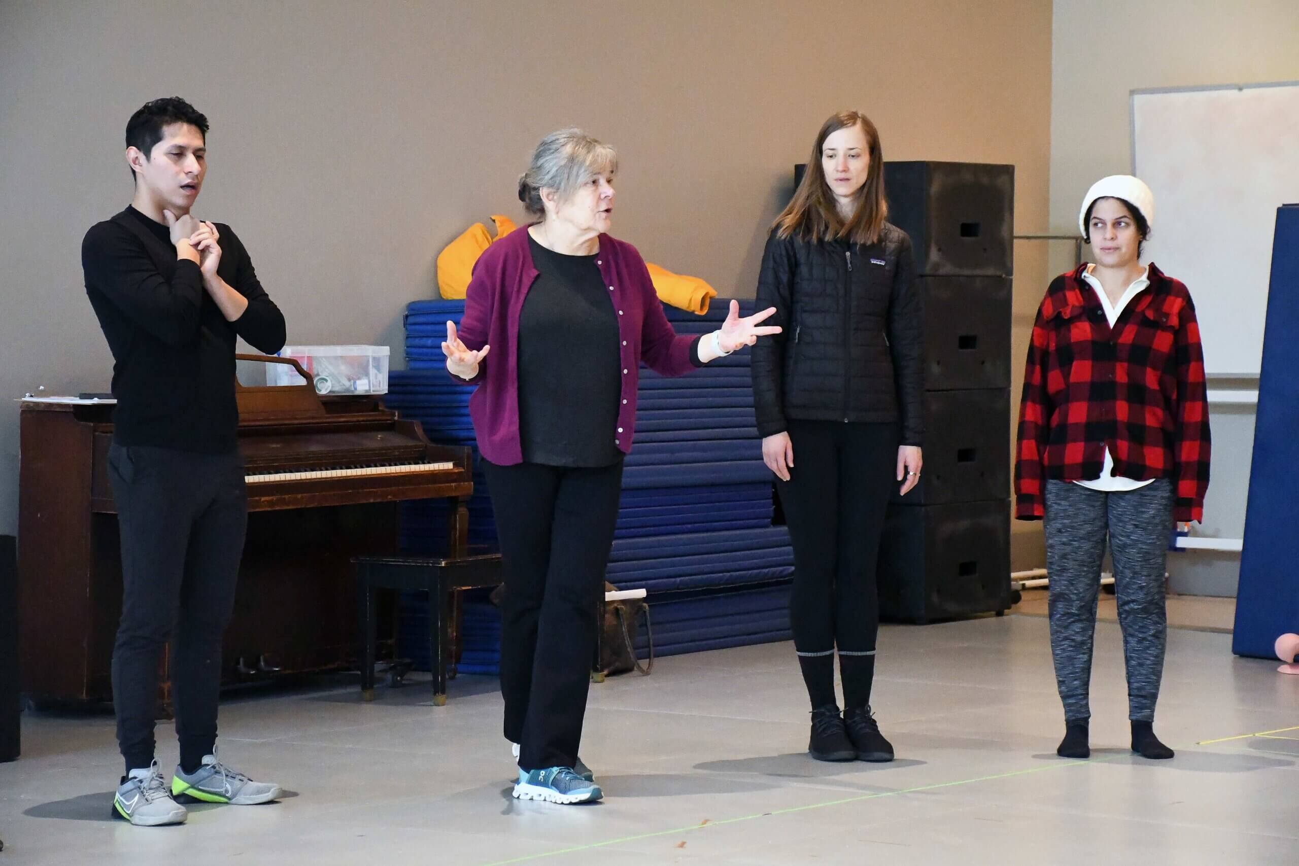 a woman in a purple sweater teaches in front of three students. THey are in a rehearsal studio