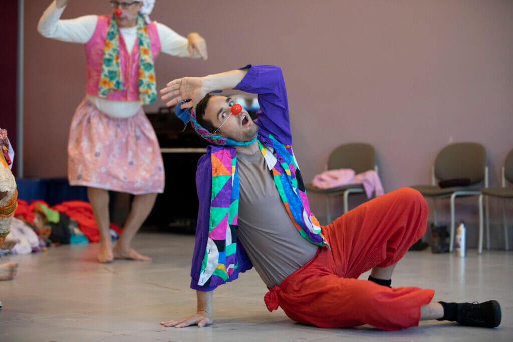 a man with a clown nose, purple jacket, and red pants sits on the florr with a hand at his head and another on the ground. He is dressed like a clown.