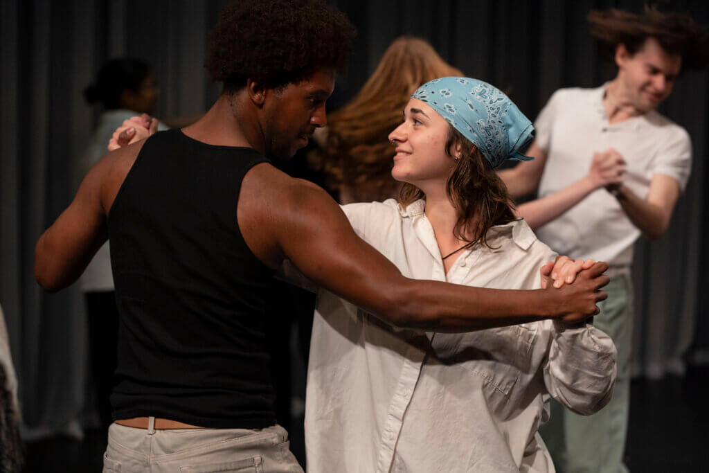 a man and woman dance. he on left is wearing a black tanktop. She on right is wearing a blue bandana on her head and a white shirt.