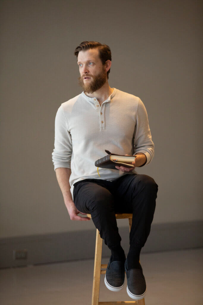 a man sits on a stool looking to the left, holding a book in his right hand