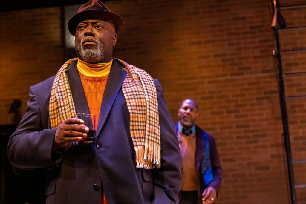 a black man with a fedora and a putple overcoat and scarf looks off to the left. he looks serious