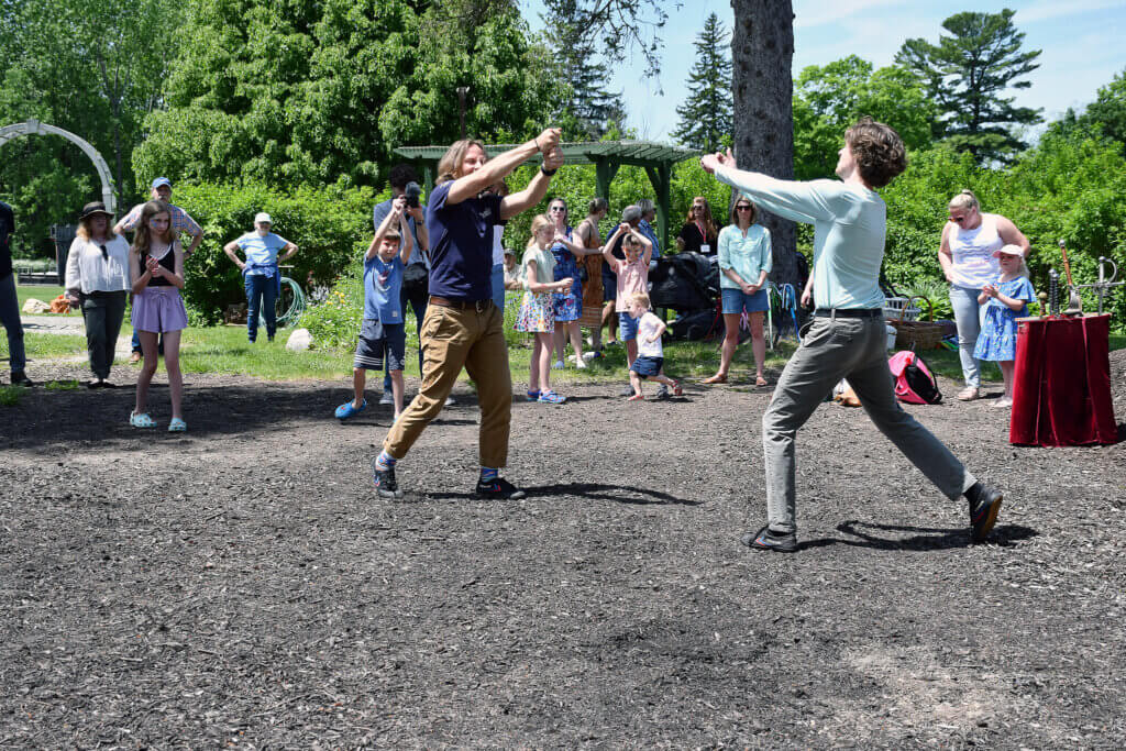 two men demonstrate stage combat outside with two wooden swords. A crowd of people watch them; it is sunny out.