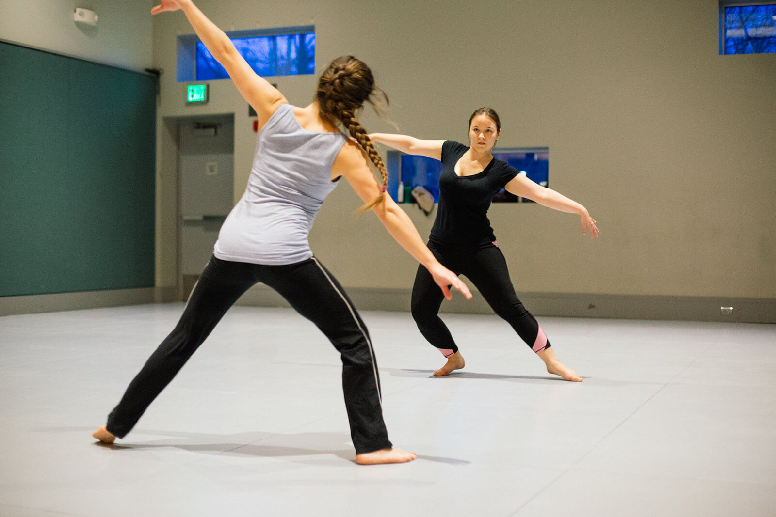 two women facing each other in a rehearsal studio with their arms outstretched and their waists bent in opposite directions.
