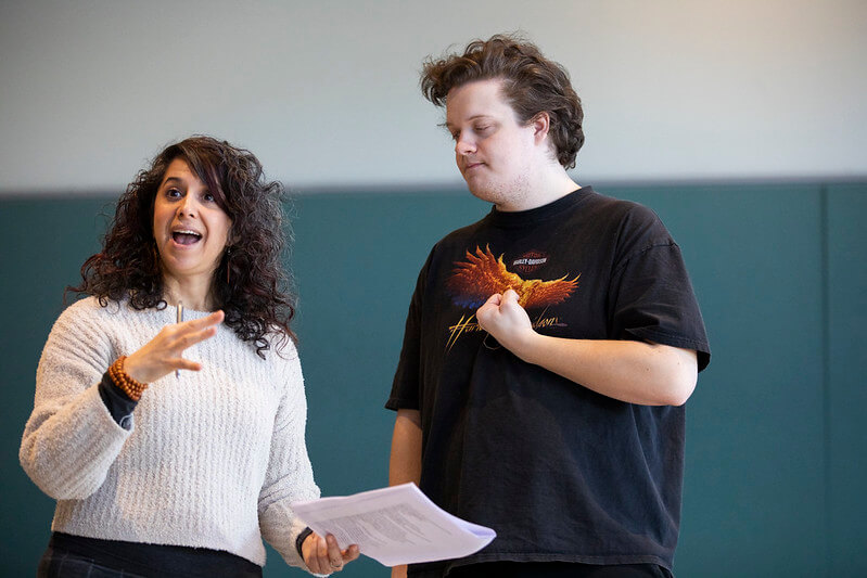 a woman with dark curly hair holds a piece of paper next to a man facing to the left wearing a black t-shirt.