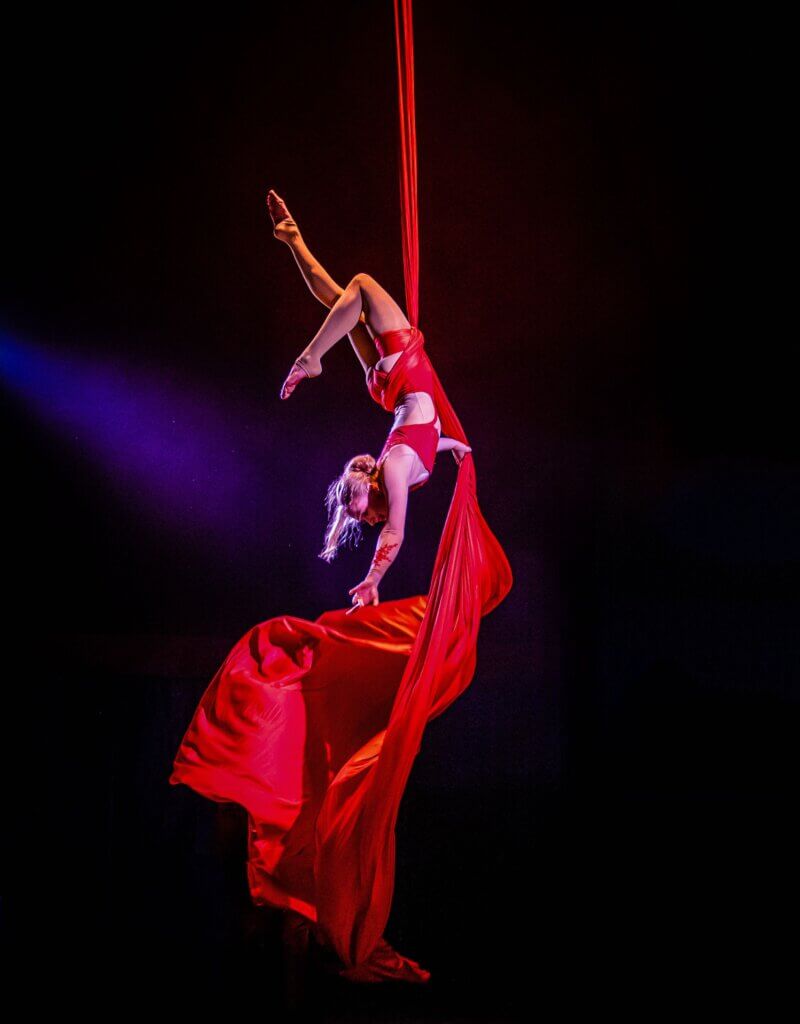 an aerialist, a woman with blonde hair wearing a red leotard, hangs upside down holding a red piece of fabric around which she is wrapped.