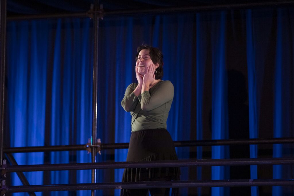 a woman on a catwalk with a bluebackground - she is wearing a green shirt, has her hands on her cheeks, and is smiling