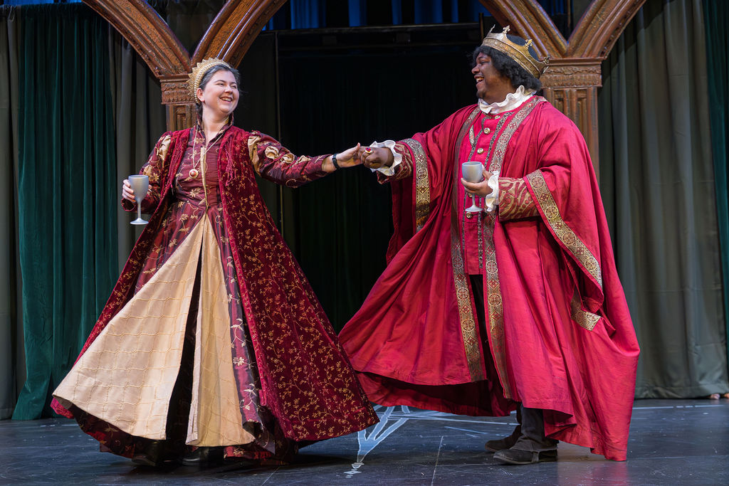a woman in a red dress and a man in a red robe hold hands and smile. Both are wearing crowns.