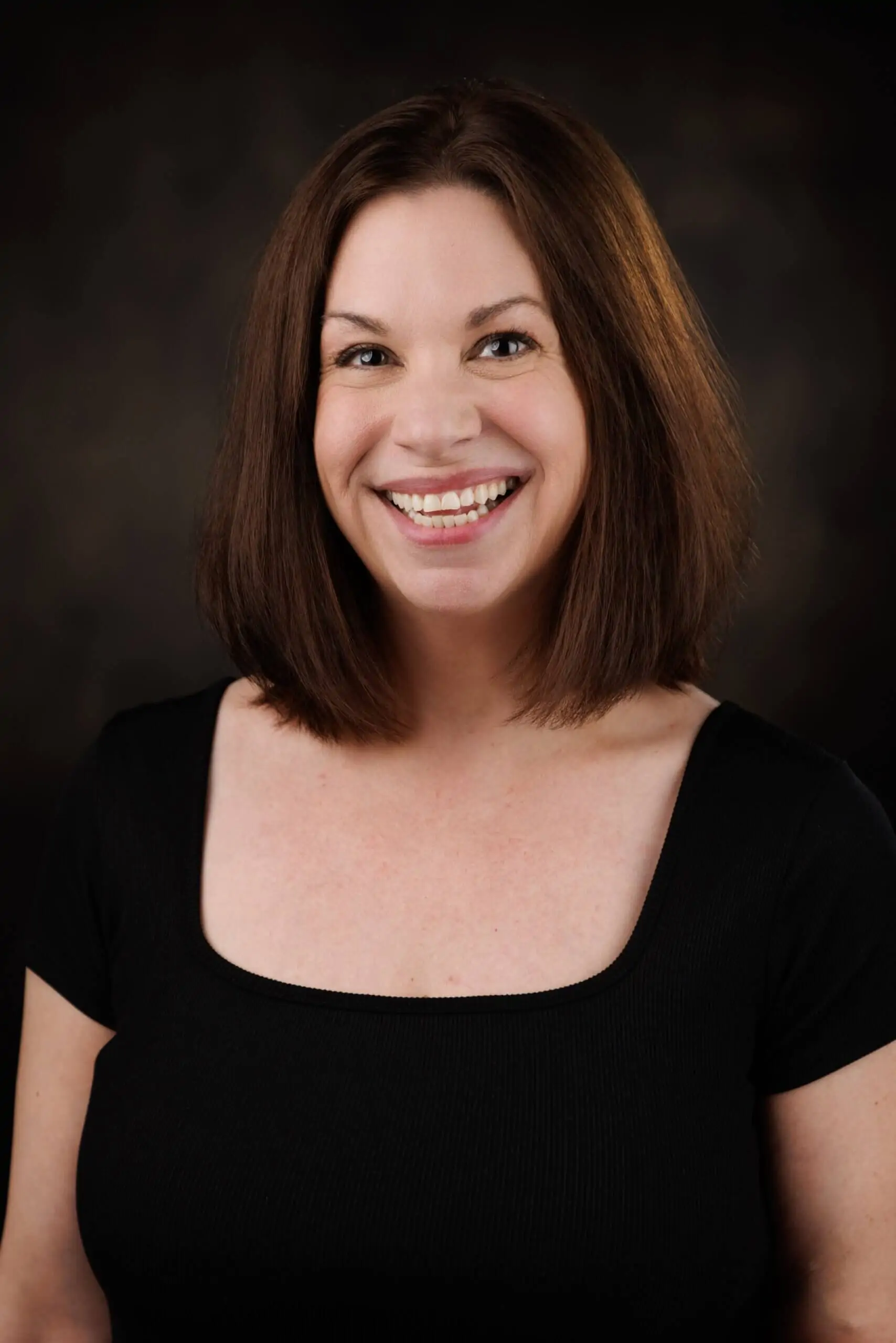 white woman with brown hair wearing a black top, smiling, in front of a brown background