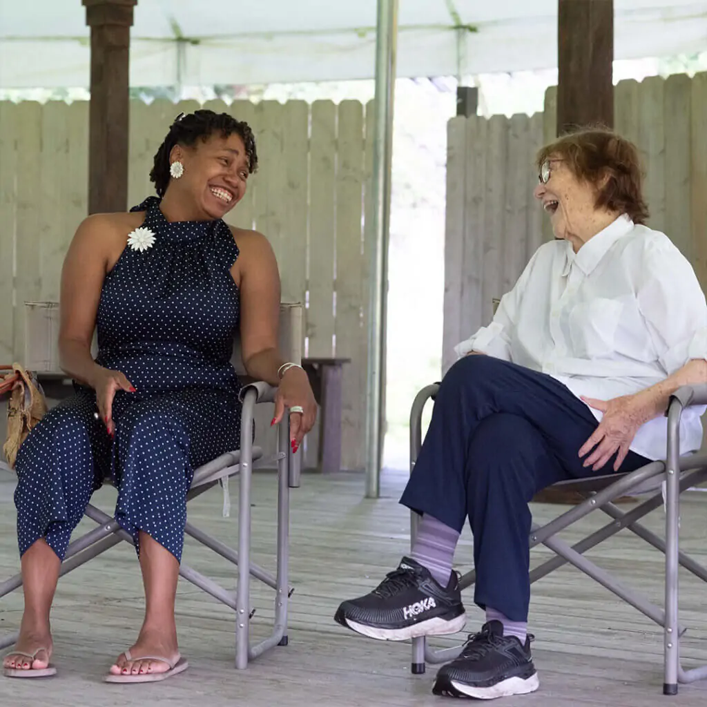two women face each other smiling, sitting on a stage in chairs. One woman on the right is older with red hair. The woman on the left is younger with black hair.