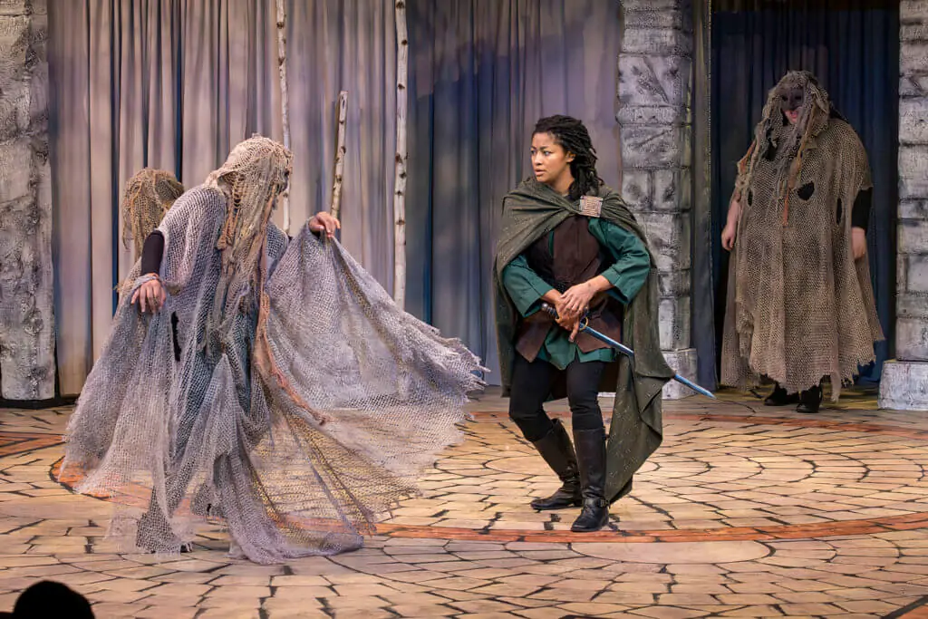 A woman at left dressed in gray gauze faces a woman in a green cape. Another woman stands at right in the same gray gauzy costume. They are performing in a staged production of Macbeth