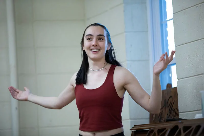 a young woman with brown hair looks to the right ans smiles. She has her arms outstretched and is wearing a red tank top. She is in a rehearsal space.
