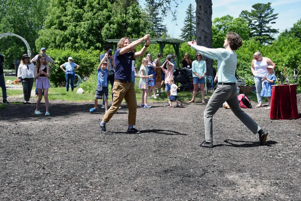 two men demonstrate stage combat outside with two wooden swords. A crowd of people watch them; it is sunny out.